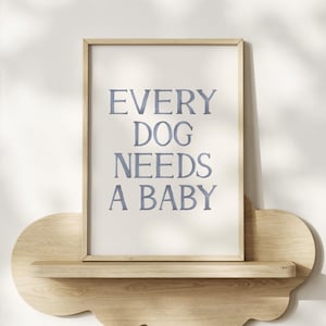 May include: A framed print with the text "EVERY DOG NEEDS A BABY" in blue lettering. The print is displayed on a light wood shelf with a decorative edge, against a white wall.
