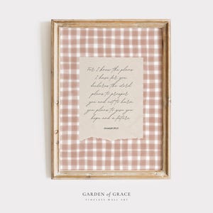 May include: Framed wall art featuring a light brown and white gingham pattern background. A torn-edged paper with handwritten text is centered. The frame is a light wood color. The text reads "For I know the plans I have for you declares the Lord..." and is from Jeremiah 29:11.