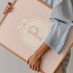 Puede incluir: Una obra de arte enmarcada con un patr&oacute;n geom&eacute;trico rosa claro. Un emblema crema con la letra "P" rosa y un lazo est&aacute; centrado en la obra. El marco es de madera clara.