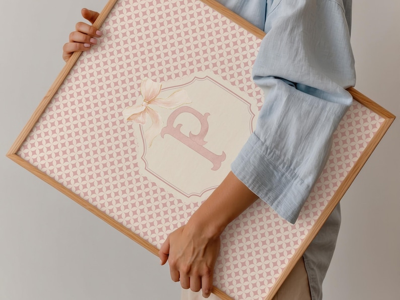 Puede incluir: Una obra de arte enmarcada con un patr&oacute;n geom&eacute;trico rosa claro. Un emblema crema con la letra "P" rosa y un lazo est&aacute; centrado en la obra. El marco es de madera clara.