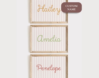 May include: Three framed prints with names: Hailey, Amelia, and Penelope. Each print has a striped background in pink and white, with the name in a different color: gold, green, and red. The frames are wooden, and a brown label says "CUSTOM NAME".