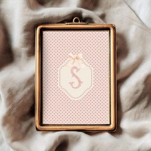 Puede incluir: Una impresi&oacute;n enmarcada con un marco dorado. La impresi&oacute;n presenta un fondo estampado en rosa y blanco con un emblema color crema en el centro. El emblema tiene una letra "S" rosa y un lazo color crema en la parte superior.