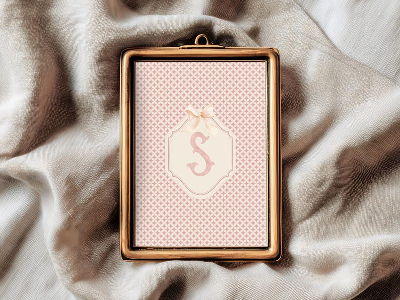 Puede incluir: Una impresi&oacute;n enmarcada con un marco dorado. La impresi&oacute;n presenta un fondo estampado en rosa y blanco con un emblema color crema en el centro. El emblema tiene una letra "S" rosa y un lazo color crema en la parte superior.