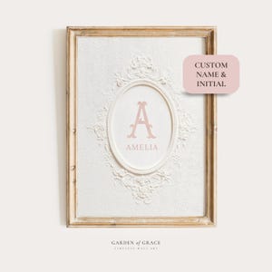 Puede incluir: Una impresión artística enmarcada con un diseño blanco en relieve y un centro ovalado. El centro muestra la letra "A" y el nombre "Amelia" en un tono rosa suave. El marco es de color dorado claro. El texto en la imagen dice "CUSTOM NAME & INITIAL" y "GARDEN OF GRACE".