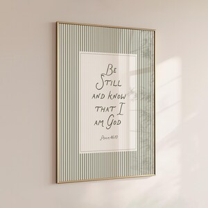 May include: Framed wall art with a gold-colored frame. The artwork features the handwritten text "Be still and know that I am God" in black ink on a cream-colored background. The frame has a striped pattern in shades of green and white.