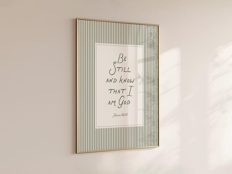 May include: Framed wall art with a gold-colored frame. The artwork features the handwritten text "Be still and know that I am God" in black ink on a cream-colored background. The frame has a striped pattern in shades of green and white.