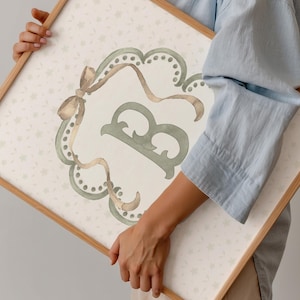 May include: A framed watercolor print featuring a decorative letter "B" in sage green. The letter is within a scalloped frame with ribbon accents in shades of brown and gold. The background has a subtle floral pattern.