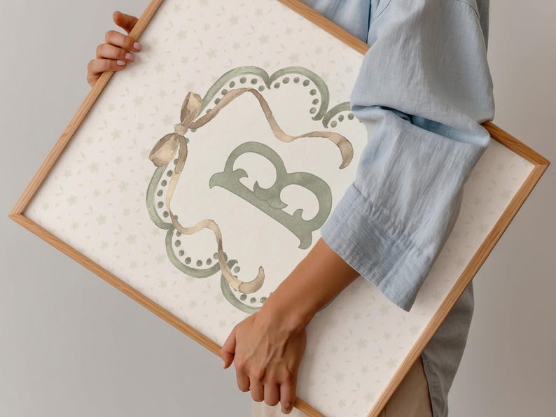 May include: A framed watercolor print featuring a decorative letter "B" in sage green. The letter is within a scalloped frame with ribbon accents in shades of brown and gold. The background has a subtle floral pattern.