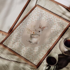 K&ouml;nnte beinhalten: Ein gerahmter Druck mit einer Aquarellillustration eines Hasen, der Blumen h&auml;lt. Der Hase ist vor einem floral gemusterten Hintergrund platziert. Eine Tasse Tee und Sahne sind sichtbar. Der Rahmen ist aus Holz.
