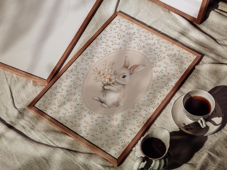 K&ouml;nnte beinhalten: Ein gerahmter Druck mit einer Aquarellillustration eines Hasen, der Blumen h&auml;lt. Der Hase ist vor einem floral gemusterten Hintergrund platziert. Eine Tasse Tee und Sahne sind sichtbar. Der Rahmen ist aus Holz.