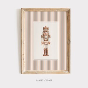 May include: Framed print featuring a watercolor illustration of a nutcracker soldier. The artwork is set against a striped background in a wooden frame. The print is labeled "GARDEN OF GRACE, TIMELESS WALL ART".