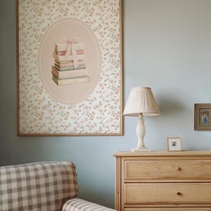 May include: A framed floral print with an oval illustration of stacked books tied with a pink bow. A cream-colored lamp with a pleated shade sits on a wooden dresser. A small framed picture and a checkered armchair are also visible.