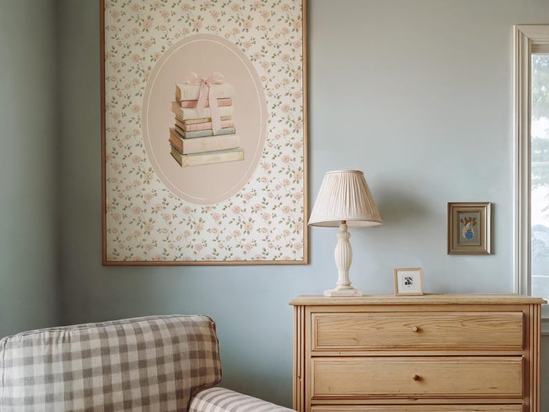 May include: A framed floral print with an oval illustration of stacked books tied with a pink bow. A cream-colored lamp with a pleated shade sits on a wooden dresser. A small framed picture and a checkered armchair are also visible.
