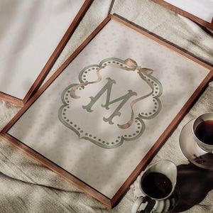 May include: A framed print featuring the letter "M" in a decorative, sage green font. The print has a light background with a subtle star pattern and is accented with a gold ribbon. A cup of coffee and creamer are in the foreground.