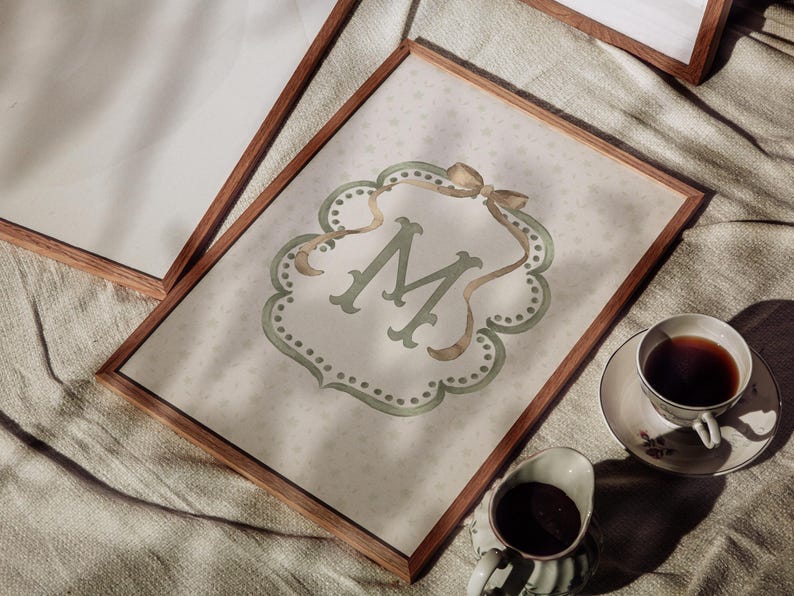 May include: A framed print featuring the letter "M" in a decorative, sage green font. The print has a light background with a subtle star pattern and is accented with a gold ribbon. A cup of coffee and creamer are in the foreground.