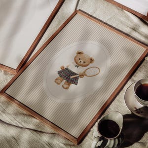 K&ouml;nnte beinhalten: Ein gerahmter Druck mit einer Aquarellillustration eines Teddyb&auml;ren in Tennisbekleidung, der einen Schl&auml;ger h&auml;lt. Der B&auml;r ist vor einem gestreiften Hintergrund in einem Oval platziert. Der Rahmen ist aus dunklem Holz. Eine Tasse Tee und Sahne sind im Vordergrund.