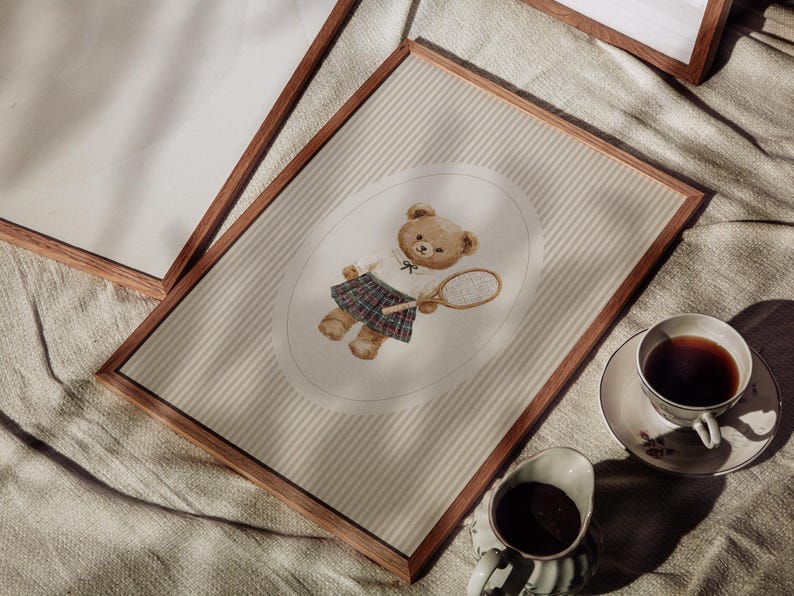 K&ouml;nnte beinhalten: Ein gerahmter Druck mit einer Aquarellillustration eines Teddyb&auml;ren in Tennisbekleidung, der einen Schl&auml;ger h&auml;lt. Der B&auml;r ist vor einem gestreiften Hintergrund in einem Oval platziert. Der Rahmen ist aus dunklem Holz. Eine Tasse Tee und Sahne sind im Vordergrund.