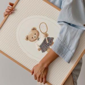 K&ouml;nnte beinhalten: Ein gerahmter Druck mit einer Aquarellillustration eines Teddyb&auml;ren in Tennisbekleidung, der einen Schl&auml;ger h&auml;lt. Der B&auml;r tr&auml;gt ein wei&szlig;es Hemd, einen karierten Rock und eine Fliege. Der Hintergrund hat beige und wei&szlig;e Streifen.