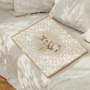 K&ouml;nnte beinhalten: Ein gerahmter Druck mit einer Aquarellillustration eines Hasen, der Blumen h&auml;lt. Der Hase ist vor einem weichen, rosa Oval platziert, umgeben von einem floralen Muster. Der Rahmen ist aus hellem Holz und liegt auf einem Bett.