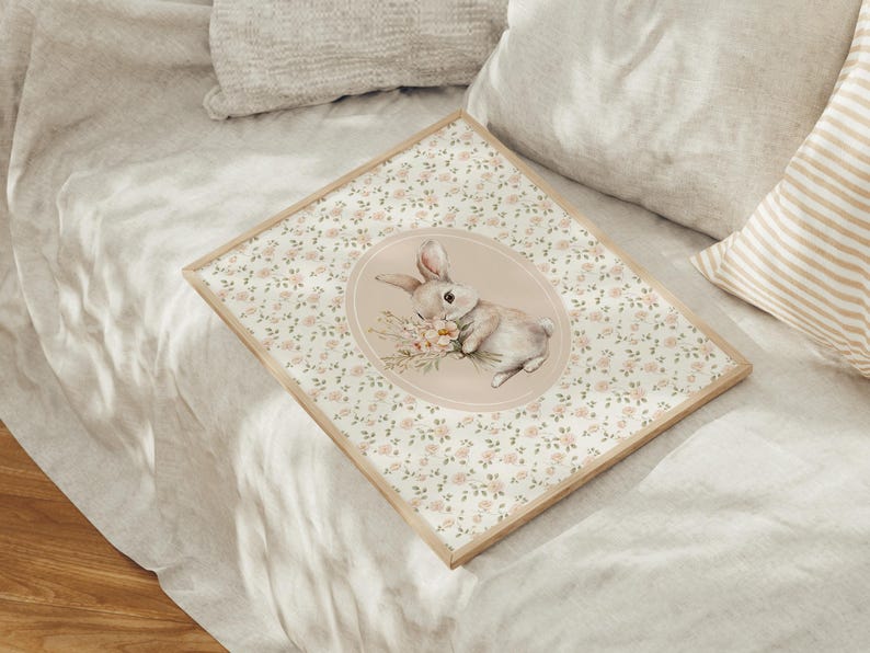 K&ouml;nnte beinhalten: Ein gerahmter Druck mit einer Aquarellillustration eines Hasen, der Blumen h&auml;lt. Der Hase ist vor einem weichen, rosa Oval platziert, umgeben von einem floralen Muster. Der Rahmen ist aus hellem Holz und liegt auf einem Bett.