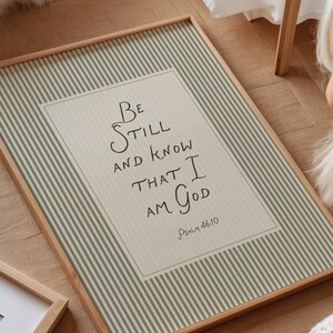 May include: A framed print with the text "Be Still and Know That I Am God" from Psalm 46:10. The print has a light beige background with a white border and a light green striped pattern. The frame is made of light brown wood.