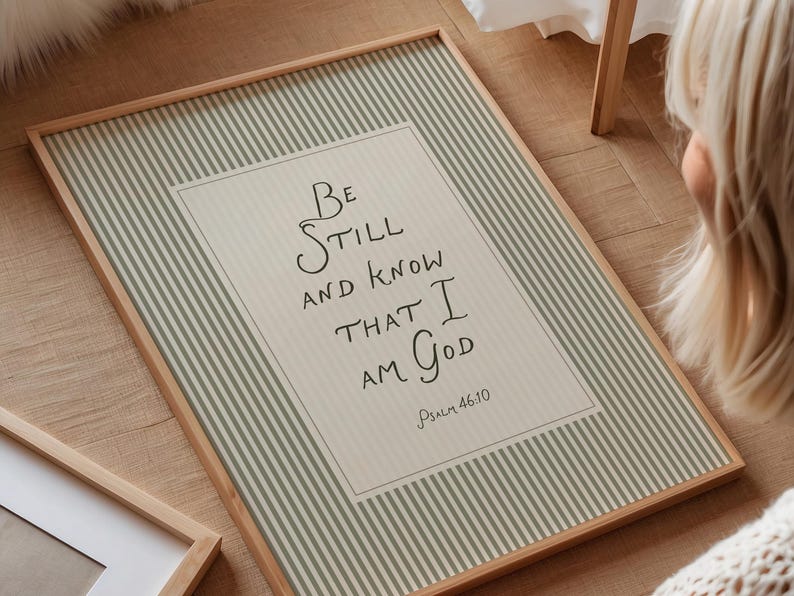 May include: A framed print with the text "Be Still and Know That I Am God" from Psalm 46:10. The print has a light beige background with a white border and a light green striped pattern. The frame is made of light brown wood.