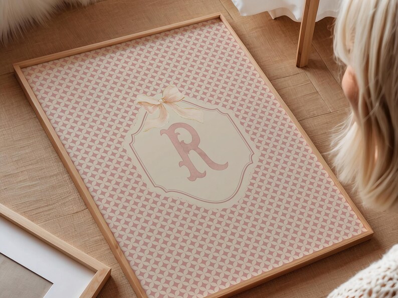 Puede incluir: Una impresi&oacute;n enmarcada con un patr&oacute;n geom&eacute;trico rosa y blanco. La impresi&oacute;n tiene un marco de madera clara y un emblema central con la letra "R" en rosa, adornado con un lazo. La est&eacute;tica general es suave y femenina.