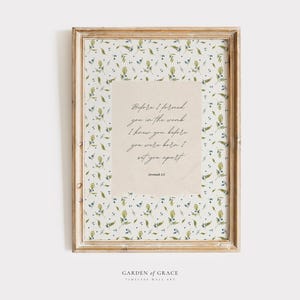 Op de afbeelding: Een ingelijst drukwerk met een witte achtergrond en een rand met een bloemenpatroon. De print bevat een citaat in cursief schrift: "Voordat ik je in de moederschoot vormde, kende ik je, voordat je geboren werd, had ik je geheiligd." Het citaat wordt toegeschreven aan Jeremia 1:5.