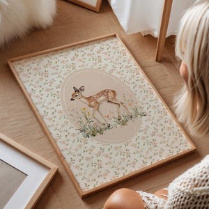 May include: A framed art print featuring a fawn illustration within an oval frame. The background is a floral pattern with pink roses on a cream-colored backdrop. The frame is made of light-colored wood.