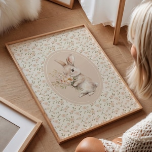 K&ouml;nnte beinhalten: Ein gerahmter Druck mit einer Aquarellillustration eines Hasen, der Blumen h&auml;lt. Der Hase ist vor einem beigen Oval platziert, umgeben von einem floralen Muster. Der Rahmen besteht aus hellem Holz, mit einer weichen, pastellfarbenen &Auml;sthetik.