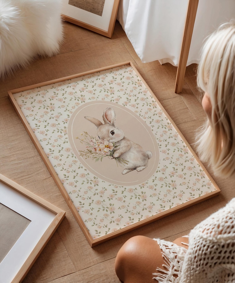 K&ouml;nnte beinhalten: Ein gerahmter Druck mit einer Aquarellillustration eines Hasen, der Blumen h&auml;lt. Der Hase ist vor einem beigen Oval platziert, umgeben von einem floralen Muster. Der Rahmen besteht aus hellem Holz, mit einer weichen, pastellfarbenen &Auml;sthetik.