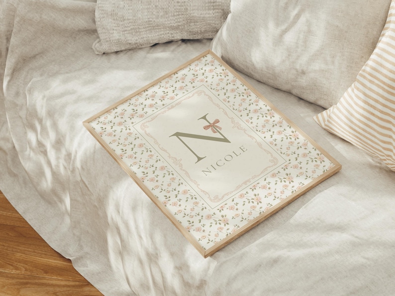 Pu&ograve; includere: Stampa incorniciata con bordo floreale e il nome "Nicole". La stampa ha una cornice in legno chiaro e un disegno centrale con la lettera "N" e un fiocco rosa. Lo sfondo &egrave; di un colore neutro.