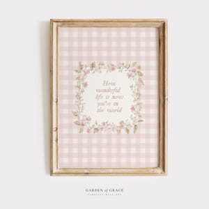 May include: Framed wall art with a pink and white gingham pattern background. The artwork features a floral wreath surrounding the text: "How wonderful life is now you're in the world." The frame is a light brown color.