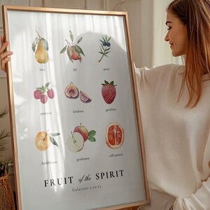 May include: A framed print with a white background and a watercolour illustration of fruits representing the fruit of the spirit. The text "FRUIT of the SPIRIT" is in bold black letters with the scripture reference "Galatians 5:22-23" below. The fruits include a pear, pomegranate, berries, figs, strawberries, apples, and an orange.