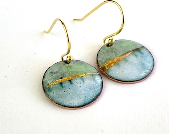 May include: A pair of round earrings with gold-colored hooks. The earrings feature a light green and blue speckled design with a gold-colored diagonal stripe. The edges of the earrings are a dark brown color.