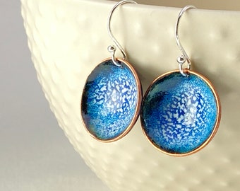 May include: A pair of handcrafted earrings with a copper rim and a blue and white speckled enamel design. The earrings have silver-colored hooks. The enamel has a speckled pattern.