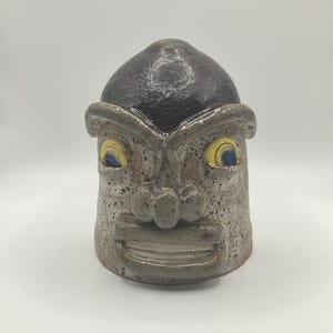 May include: A ceramic sculpture shaped like a head with a dark brown, smooth top and a speckled, light gray base. The face features prominent, protruding eyebrows, a wide nose, and a mouth with visible teeth. The eyes are yellow with blue pupils.