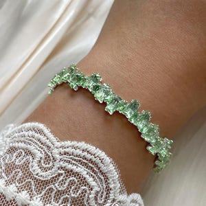 Könnte beinhalten: Ein hellgrünes Armband mit rechteckigen, facettierten Perlen. Das Armband wird an einem Handgelenk getragen, darunter ist ein weißer Spitzensaum zu sehen. Der Text "Chain Reverie" befindet sich am unteren Bildrand.