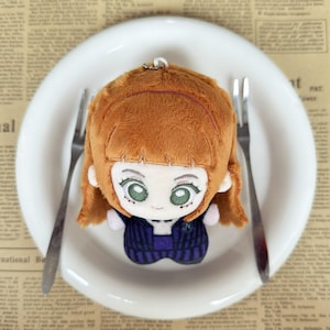 May include: A plush doll with ginger hair, green eyes, and a dark blue and purple outfit sits on a white plate with two forks. The doll has a key chain attached to the top of its head. The background is a newspaper.
