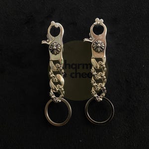May include: Two silver-toned metal keychains featuring a spring-loaded lobster clasp with a circular floral emblem, a short curb chain with decorative cross-shaped links, and a round split ring at the bottom for holding keys or other attachments.