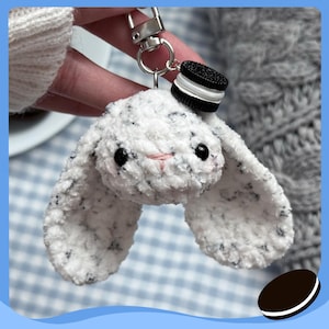 Könnte beinhalten: Ein handgefertigter, weiß-schwarz gesprenkelter Hasen-Schlüsselanhänger mit schwarzen Augen und einer rosa Nase. Der Hase ist an einem silbernen Schlüsselanhänger mit einem kleinen Keks-Anhänger befestigt.