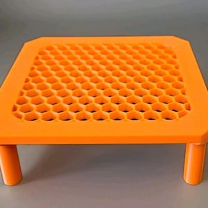 May include: Bright orange, square-shaped platform with a honeycomb pattern on top. The platform is supported by four short, cylindrical legs. The design is modern and geometric.