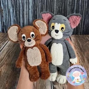 Könnte beinhalten: Ein braunes gehäkeltes Maus-Plüschtier und ein graues gehäkeltes Katzen-Plüschtier. Die Maus hat große Ohren und einen weißen Bauch. Die Katze hat rosa Ohren und einen weißen Bauch. Die Spielzeuge liegen auf einer Holzoberfläche.