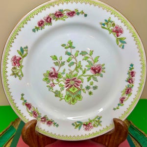 May include: White ceramic plate with a floral design featuring pink and green flowers. The plate has a gold rim and a green border with a scalloped edge. The plate is displayed on a wooden stand.