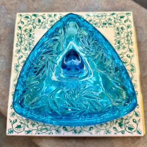 May include: A triangular, turquoise-colored glass dish with intricate leaf-like patterns. The dish has a small, rounded indentation in the center and sits on a patterned, off-white surface with green floral designs. The glass is transparent.