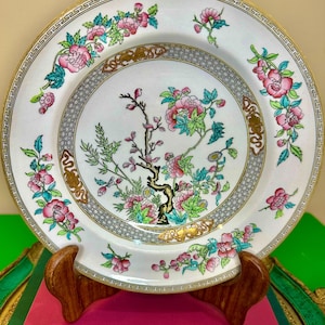 May include: A white decorative plate with a floral design. The plate features pink flowers, green leaves, and gold accents. The plate is displayed on a wooden stand, showcasing its intricate details and vintage charm.