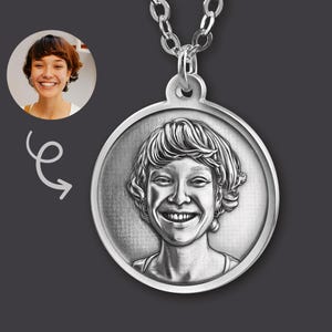 Op de afbeelding: Een ronde, zilverkleurige hanger aan een bijpassende ketting, voorzien van een gedetailleerd, gegraveerd portret van een lachend persoon met kort haar. De hanger toont een etsachtige afbeelding met een hoog contrast.