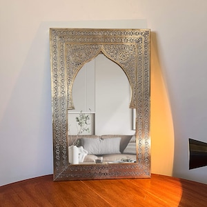 May include: A rectangular mirror with an arched top and an ornate gold-coloured frame. The frame features intricate patterns and designs. The mirror reflects a room with a sofa, cushions, and decorative items. The mirror is placed on a wooden surface.