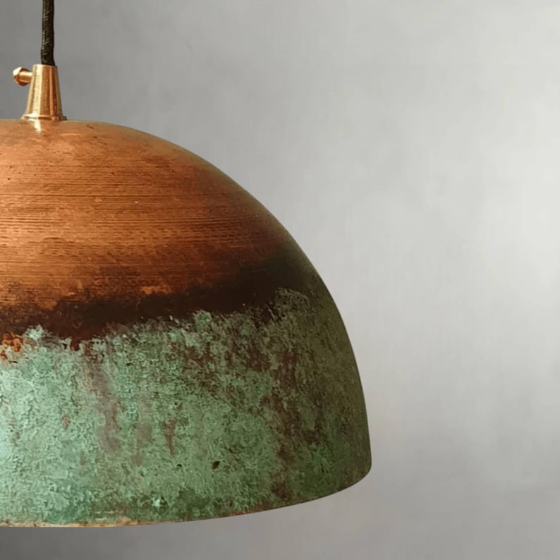 May include: A copper pendant light with a dome-shaped shade. The top of the shade is a warm copper color, transitioning to a darker brown band, and then to a textured, green patina at the bottom. The fixture has a brass-colored top and a black cord.