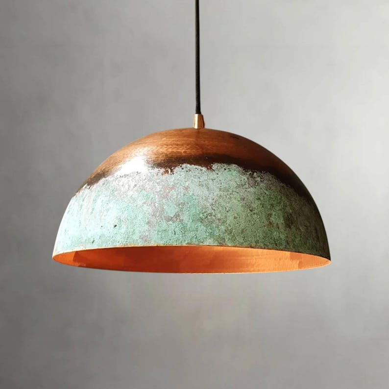 May include: A copper pendant light with a dome-shaped shade. The top of the shade is a dark brown color, transitioning to a turquoise green in the middle, and a copper interior. The light hangs from a black cord.